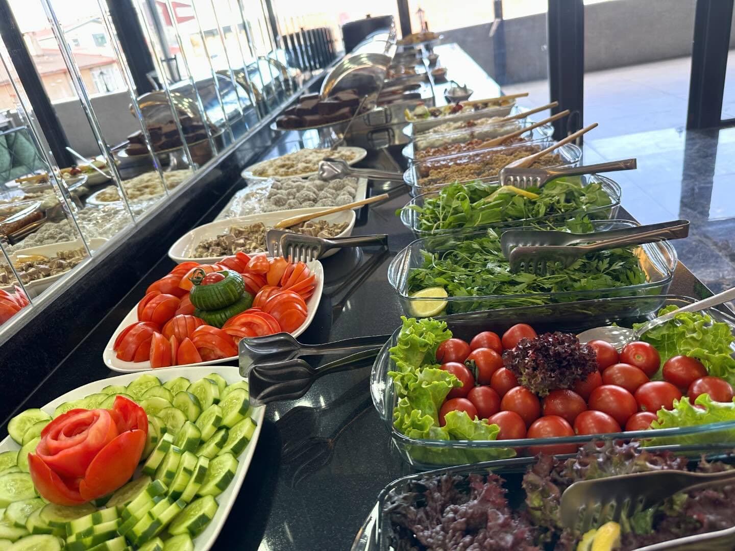 Cam tabaklarda taze salatalar; arka planda mevsim sebzeleri ve yeşillikler